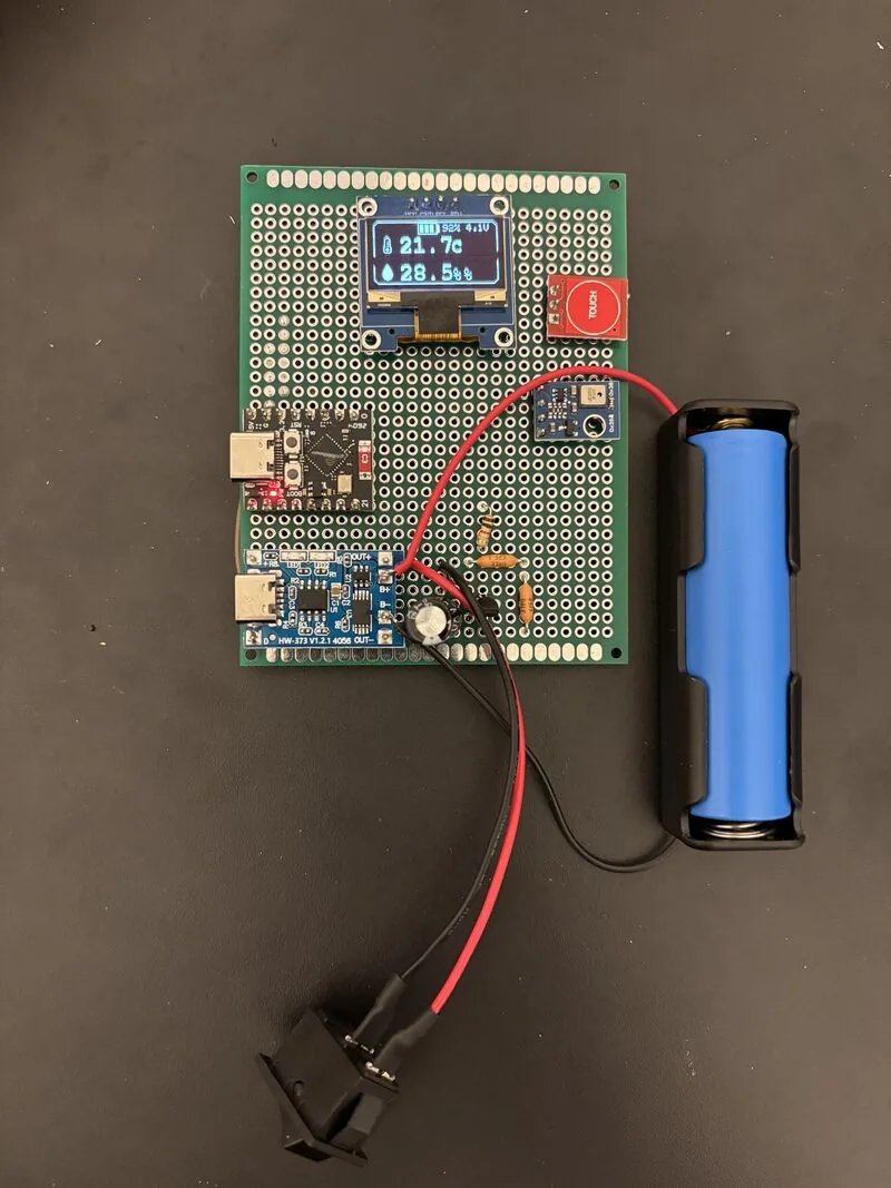 Battery-powered weather station prototype: ESP32-C3 Super Mini, OLED showing 21.7°C / 28.5% humidity, TP4056 charging board, capacitive touch button, and an 18650 cell holder, all on a perfboard